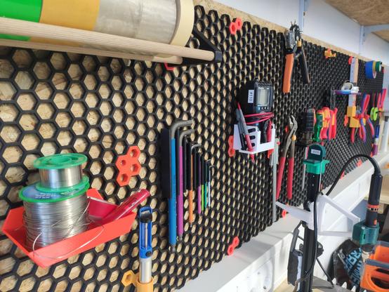 An einer Wand (OSB Platten) hängt ein Sechskant-Honigwabenraster in schwarz. An diesem können einzelne Werkzeuge befestigt werden. Dies geschieht mit 3D gedruckten Halterungen in orange. Unter anderem Multimeter, Zangen, Lötpumpe und mehr. Darunter kann man den weißen Brüstungskanal erkennen.